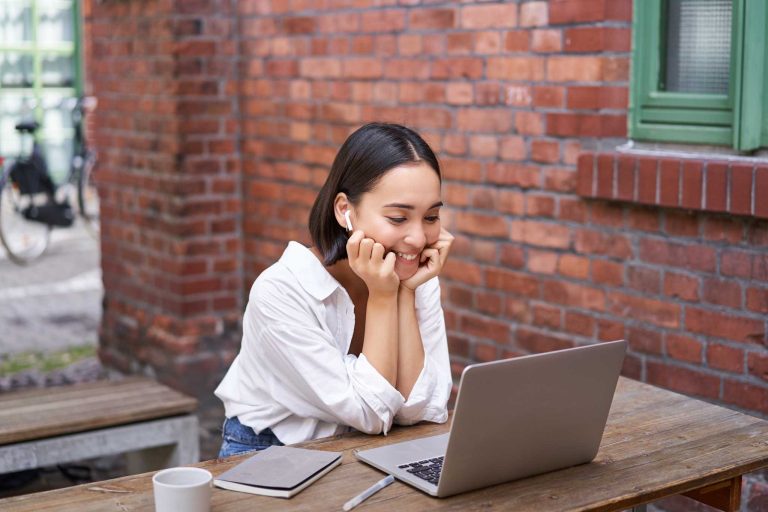 Happy asian girl taking Journey on Demand on laptop, doing homework outdoor in coworking space, smiling and working, drinking coffee.